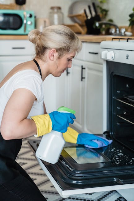 Close-up of a person's hands wearing bright yellow rubber gloves as they clean a white wall or tiled surface with a blue sponge. The other hand holds a clear spray bottle filled with cleaning solution