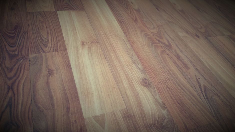 Close-up of a clean, laminated wooden floor with a smooth, glossy surface reflecting light, showing natural wood grain patterns in light beige and tan hues. The floor appears well-maintained, free of